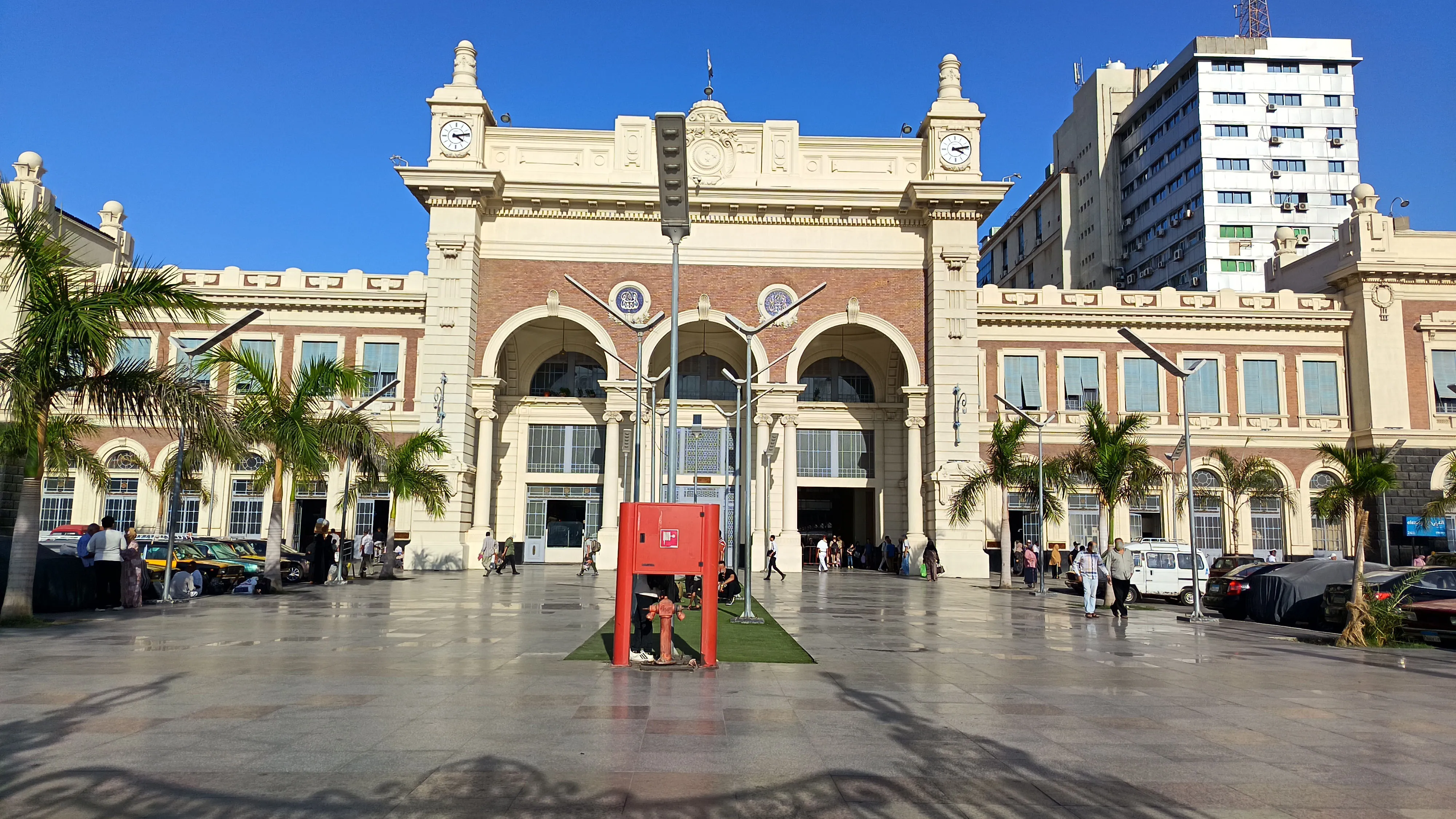 Exterior facade of Misr Train Station in Alexandria, Egypt, showing its historic colonial-era architecture with arched entrances, twin clock towers, brick detailing, and palm trees lining the forecourt on a clear sunny day.