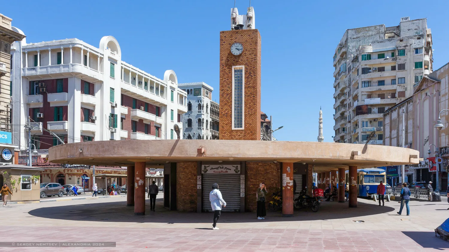 Raml Station square in Alexandria with its brick clock tower, round concrete roof and tall apartment buildings under a blue sky